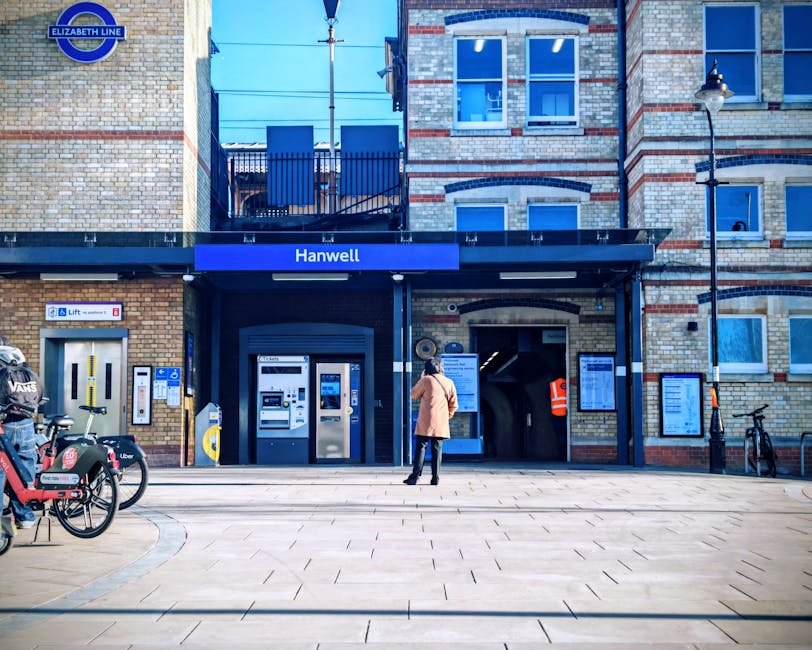 A woman standing outside the Hanwell station entrance, which features a dark blue canopy with the station name in white. The station building is constructed from light-colored brick with red accents a