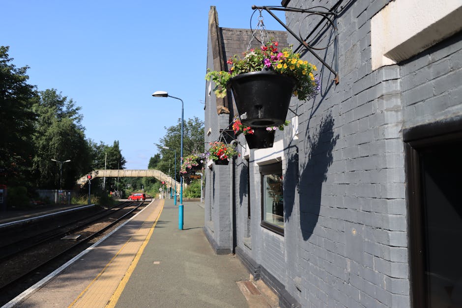 A hanging black flower basket filled with pink, yellow, purple, and white blossoms, suspended from a metal bracket on a grey brick building on a clear, sunny day. Additional smaller flower baskets wit
