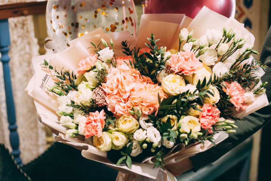 A vibrant floral bouquet featuring soft peach and white roses, complemented by white lisianthus, pink carnations, and sprigs of green foliage, arranged in an elegant style. The flowers are wrapped in