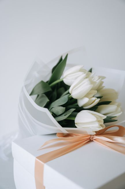A bouquet of fresh white tulips with green stems and leaves, elegantly arranged and wrapped in white tissue paper, resting atop a white gift box tied with a peach-colored ribbon. The bouquet is positi