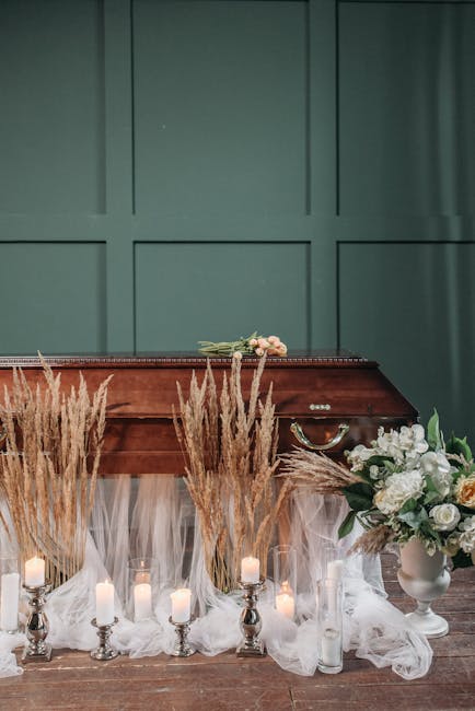 A floral arrangement featuring a large bouquet of white roses, peonies, and greenery in a white ceramic vase, placed on a wooden surface against a green paneled wall. Surrounding the arrangement are t