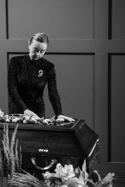 A woman dressed in a dark, textured outfit with a decorative brooch arranges a floral funeral display on a black table. She is placing light-colored flowers, possibly roses or lilies, onto the arrange