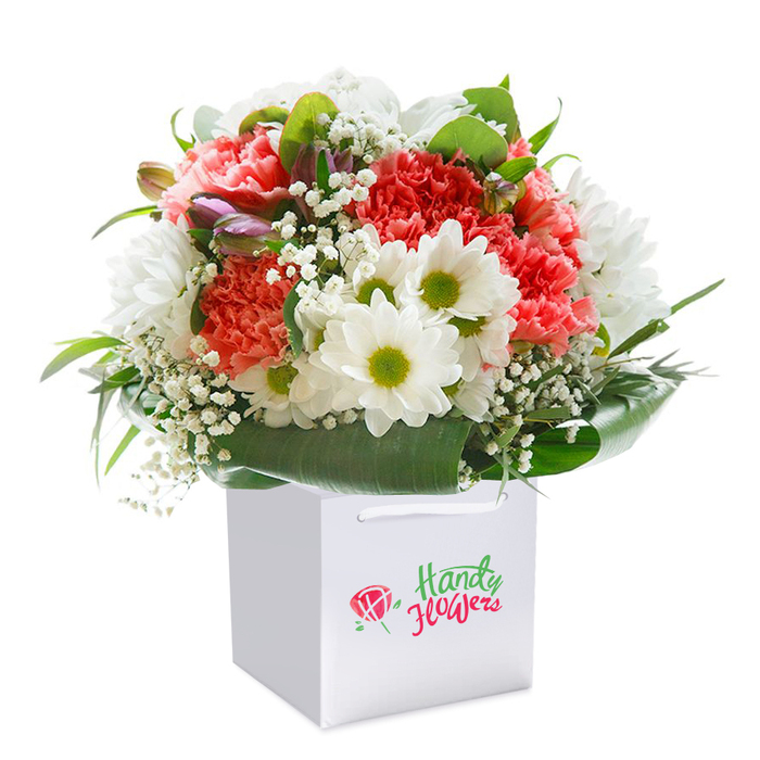 This floral masterpiece, inspired by Harlow's celebrated gardens and green spaces, showcases a sophisticated mix of colour, texture, and fresh design. Generous coral and peach carnations with their signature abundant, ruffled petals form the heart of the bouquet, radiating energy and tenderness. Bright white daisies, each with vivid yellow-green centers, add cheerful charm and visual sparkle. Airy clusters of white baby's breath thread through the display, imbuing the bouquet with lightness and movement, while purple-tipped closed buds peep softly from between the blooms, introducing subtle intrigue. Wide, deep green leaves form an elegant collar, grounding the arrangement with natural grace reminiscent of the riverside walks near The Water Gardens. Placed upright in a spotless square white gift bag, complete with comfortable ribbon handles, this bouquet is ready to make its way to any doorstep in Harlow, from New Hall to Templefields. Photographed on a crisp, modern white background, every nuance-from the glossy foliage to the fresh textures of the petals-can be appreciated. The bouquet's thoughtful composition speaks to both the skill of local florists and the ever-present beauty of Essex's landscapes, perfectly suited for birthdays, anniversaries, or spontaneous acts of kindness.
